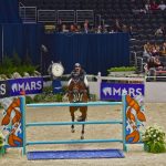 Cavaleiro conduz cavalo baio durante um salto sobre obstáculo em uma arena de hipismo.