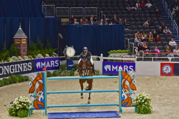 Cavaleiro conduz cavalo baio durante um salto sobre obstáculo em uma arena de hipismo.