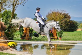 Cavaleiro e cavalo saltam sobre um obstáculo de água durante uma prova equestre.